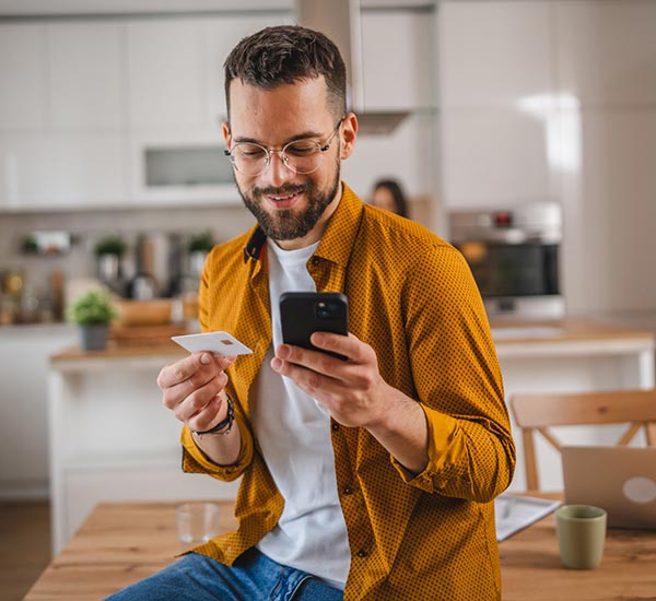 Man looking at his phone and holding a credit card in the kitchen at home.